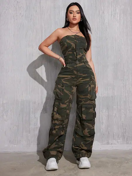 A woman with long, dark hair poses confidently in a strapless camouflage jumpsuit with a fitted bodice and cargo-style pants. The outfit features multiple pockets and a front zipper. She pairs it with white sneakers and minimal accessories, standing against a textured gray wall with a strong stance.