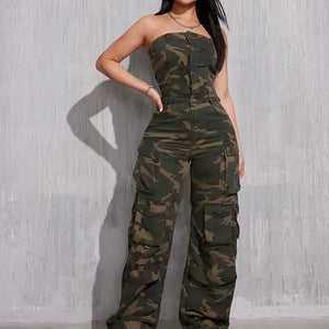 A woman with long, dark hair poses confidently in a strapless camouflage jumpsuit with a fitted bodice and cargo-style pants. The outfit features multiple pockets and a front zipper. She pairs it with white sneakers and minimal accessories, standing against a textured gray wall with a strong stance.