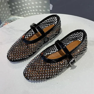 A pair of stylish black mesh Mary Jane flats adorned with shimmering rhinestones is displayed on a white round table. The shoes feature a glossy black strap with a silver buckle, a pointed toe design, and a tan insole. The background has soft lighting reflections, adding to the luxurious aesthetic.