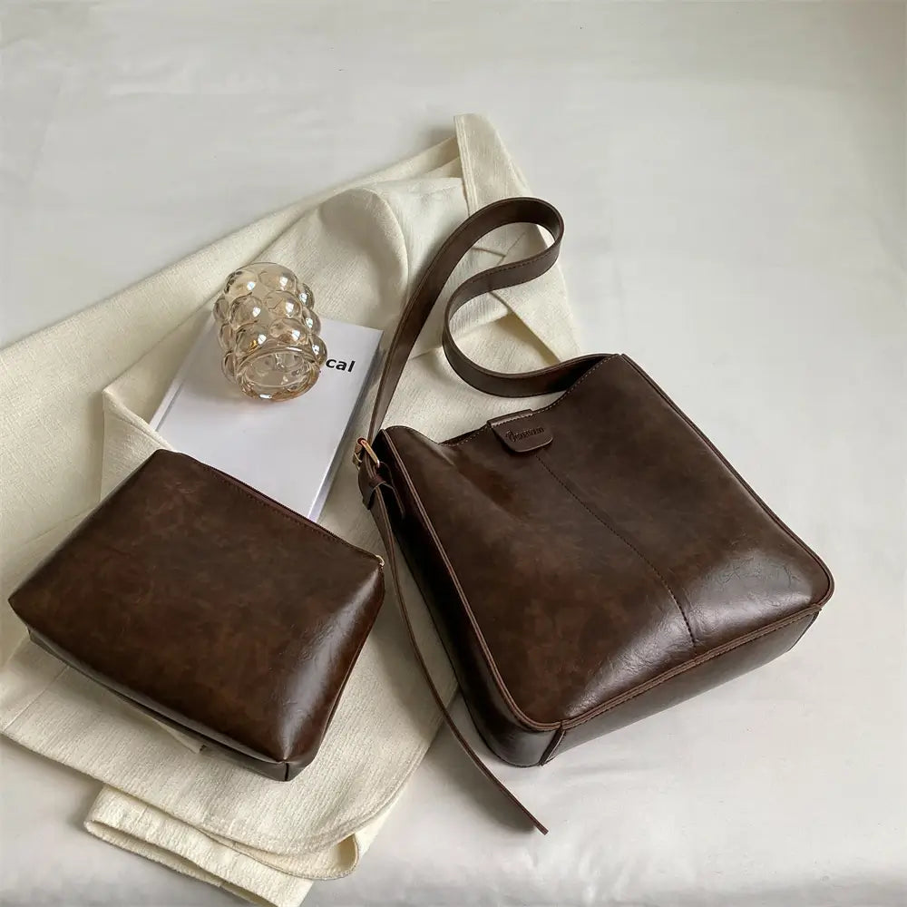 A stylish brown leather crossbody bag with a matching pouch is elegantly displayed on a white fabric surface. A clear glass with gold accents and a white magazine are placed nearby, creating a minimalistic and sophisticated aesthetic. The bag has a sleek design with a long adjustable strap and subtle branding on the front flap.